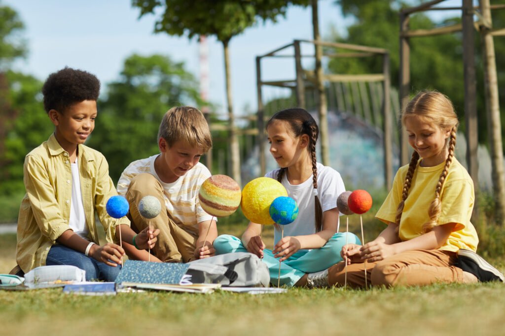Full length portrait of multi-ethnic group o children sitting on green grass and holding model planets while enjoying outdoor astronomy lesson in sunlight